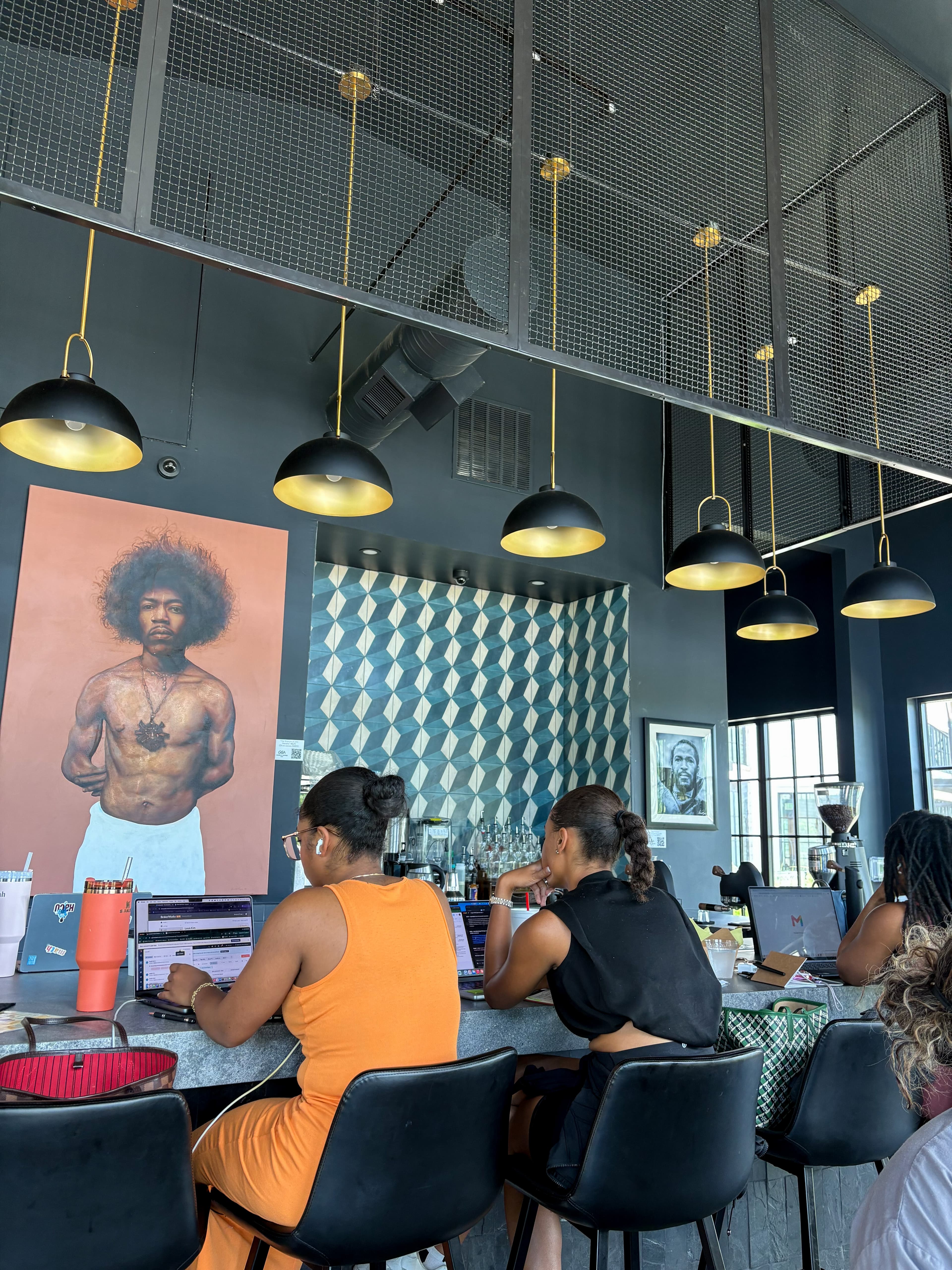 Interior of a coffee shop with people working on laptops beneath hanging lights, artwork, and tall windows.